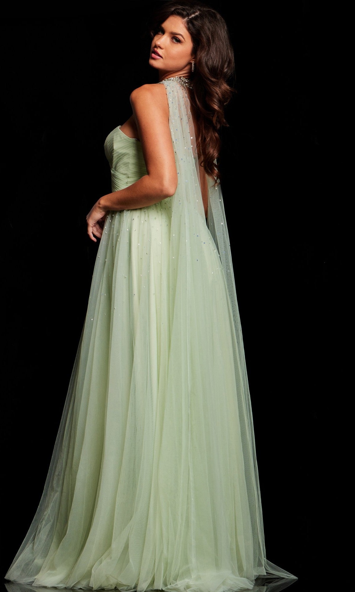 A woman in the Jovani 39307 long prom dress, light green and strapless with a sheer flowing cape, stands against a black background, glancing over her shoulder.